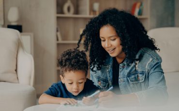 Pretty mixed race mom and little kid son focused on drawing with colored felt tip pens, playing in living room at home, spending leisure time together on weekend. Family and loving parents concept